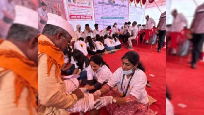 A group of healthcare volunteers, including women wearing white t-shirts and gloves, provide foot and leg massage and medical aid to Warkaris (devotees) during the Ashadhi Wari pilgrimage in Maharashtra. One elderly Warkari dressed in traditional attire with a saffron scarf receives treatment, while others wait in line inside a red-carpeted medical tent. A large banner with health initiative details is visible in the background.